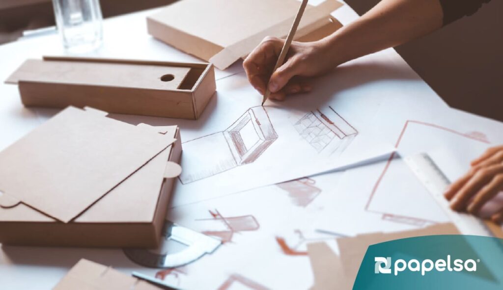 Una persona diseña y dibuja bocetos de empaques de cartón sobre una mesa, rodeada de plantillas y cajas en proceso de diseño.